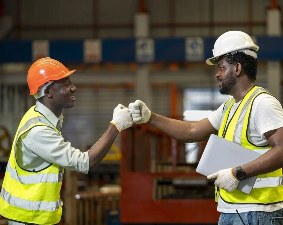 Two construction workers wearing hard hats, safety vests, and gloves fist bumping in an industrial warehouse setting during STCW safety training