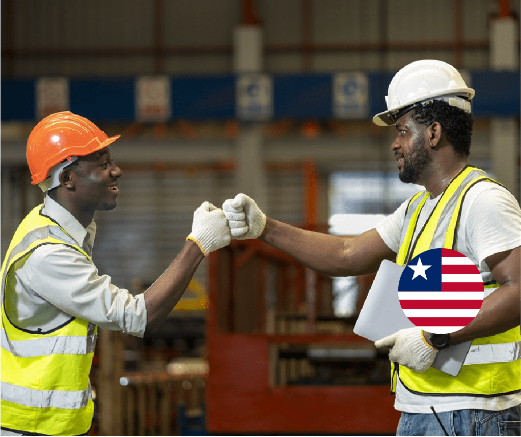 Two construction workers in safety gear fist bumping at warehouse, wearing hard hats, reflective vests, and gloves with Liberia flag badge visible