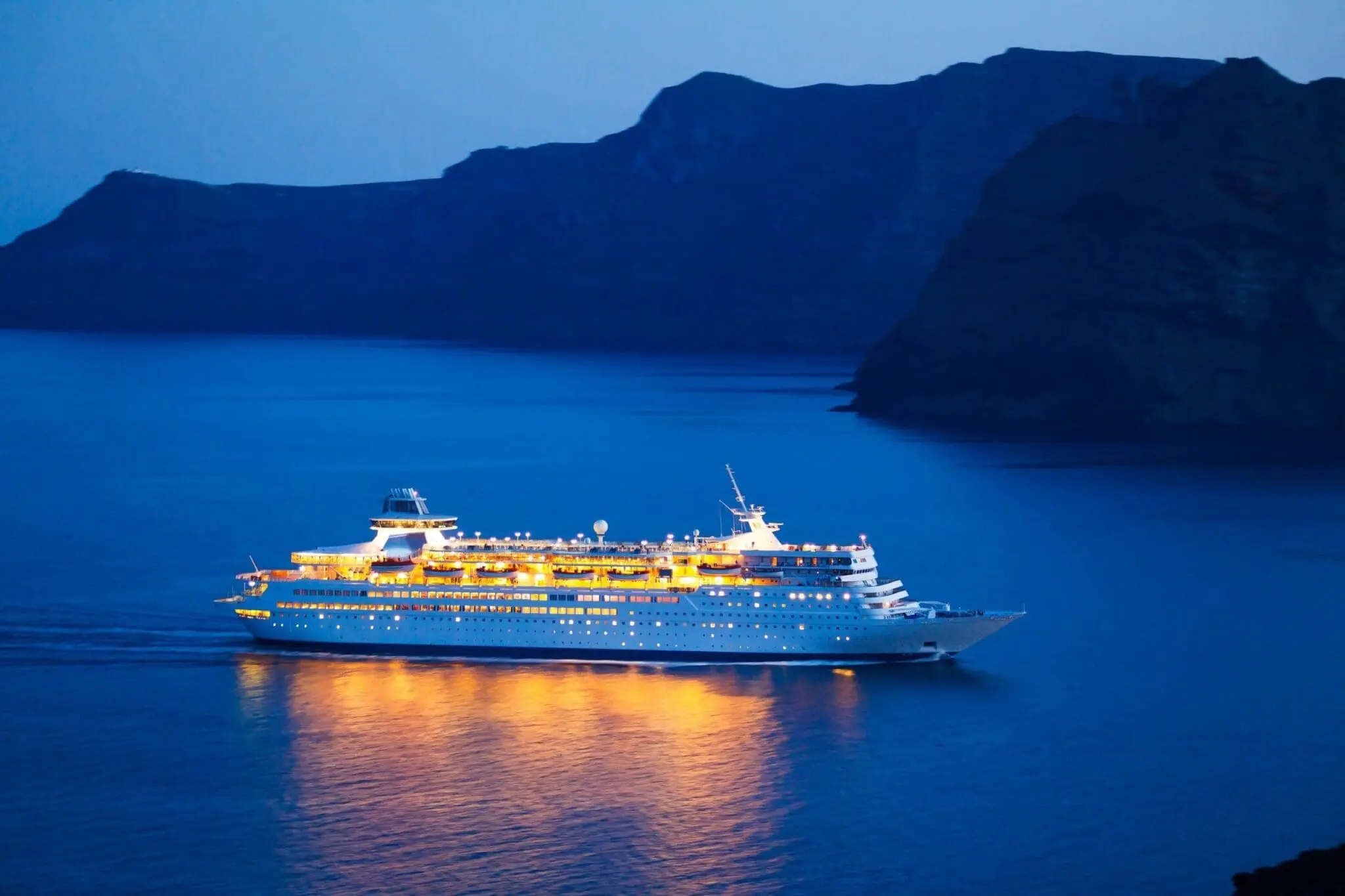 Large cruise ship with golden deck lights anchored in calm blue waters surrounded by dark rocky cliffs at twilight