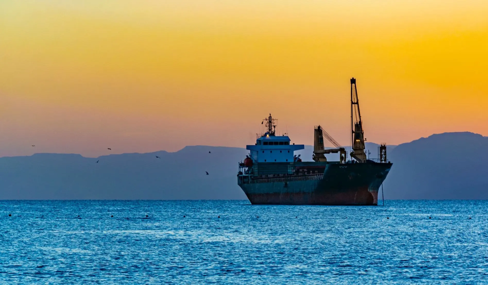Large cargo ship with blue superstructure and dual cranes anchored in bay during golden sunset with mountains in background