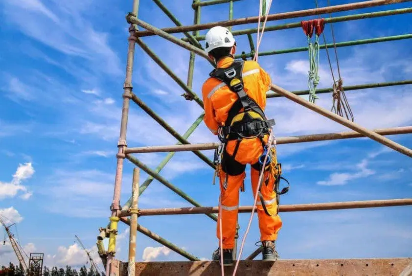 Construction worker wearing orange safety suit, white helmet, and harness secured with rope while working at height on metal scaffolding structure