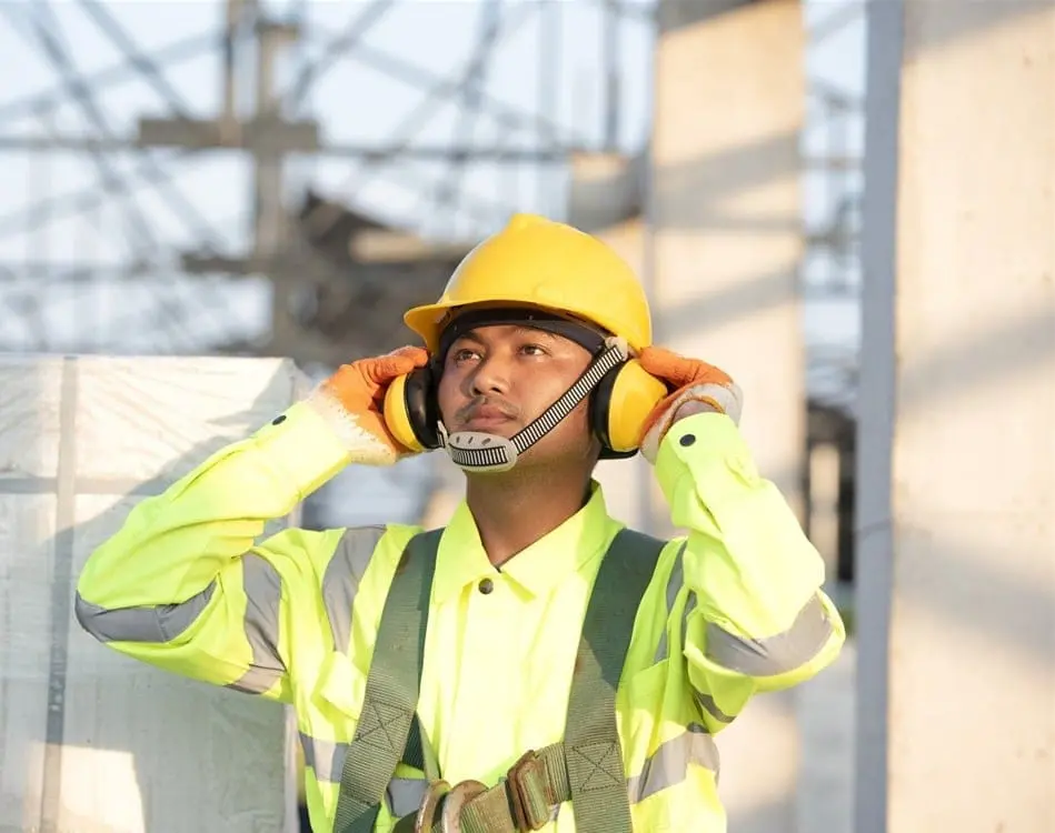 Construction worker in yellow hard hat and high-visibility vest adjusting ear defenders on a job site, demonstrating proper noise awareness safety equipment