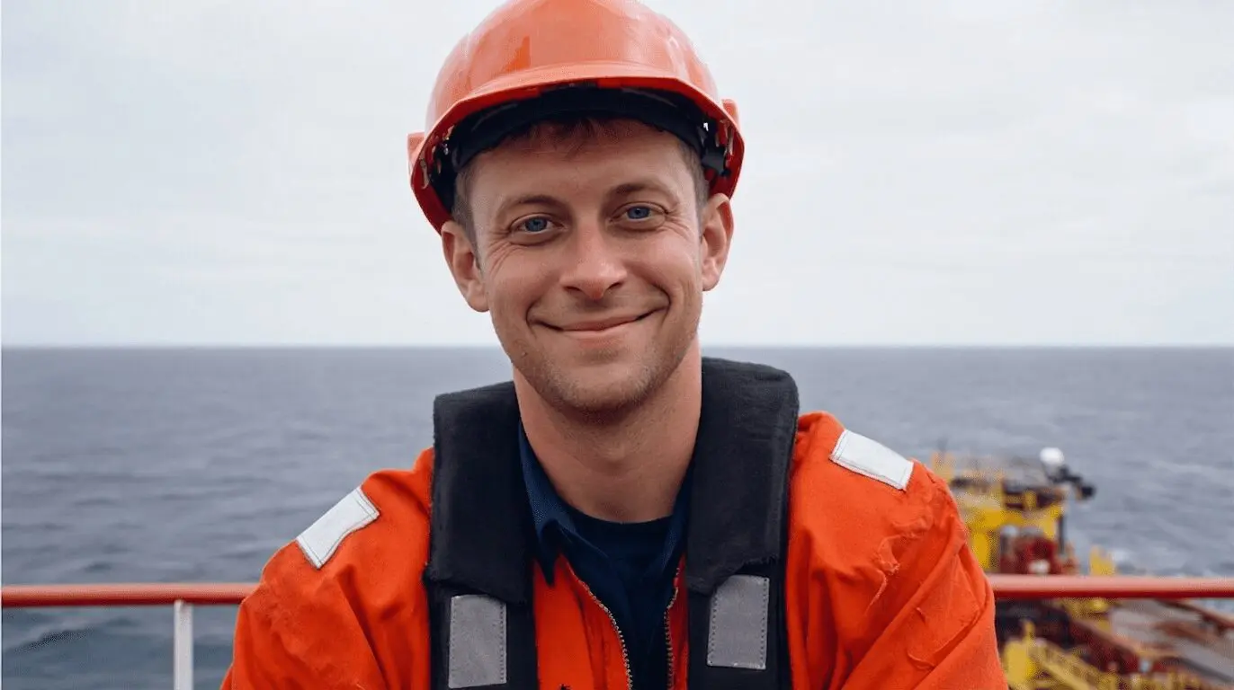 Smiling offshore worker wearing red hard hat and orange safety vest on ship deck with calm sea background