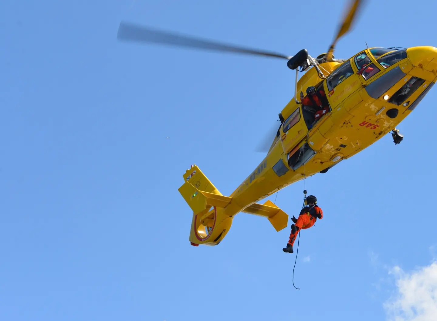 Yellow rescue helicopter with crew member performing emergency evacuation lowering operation against clear blue sky