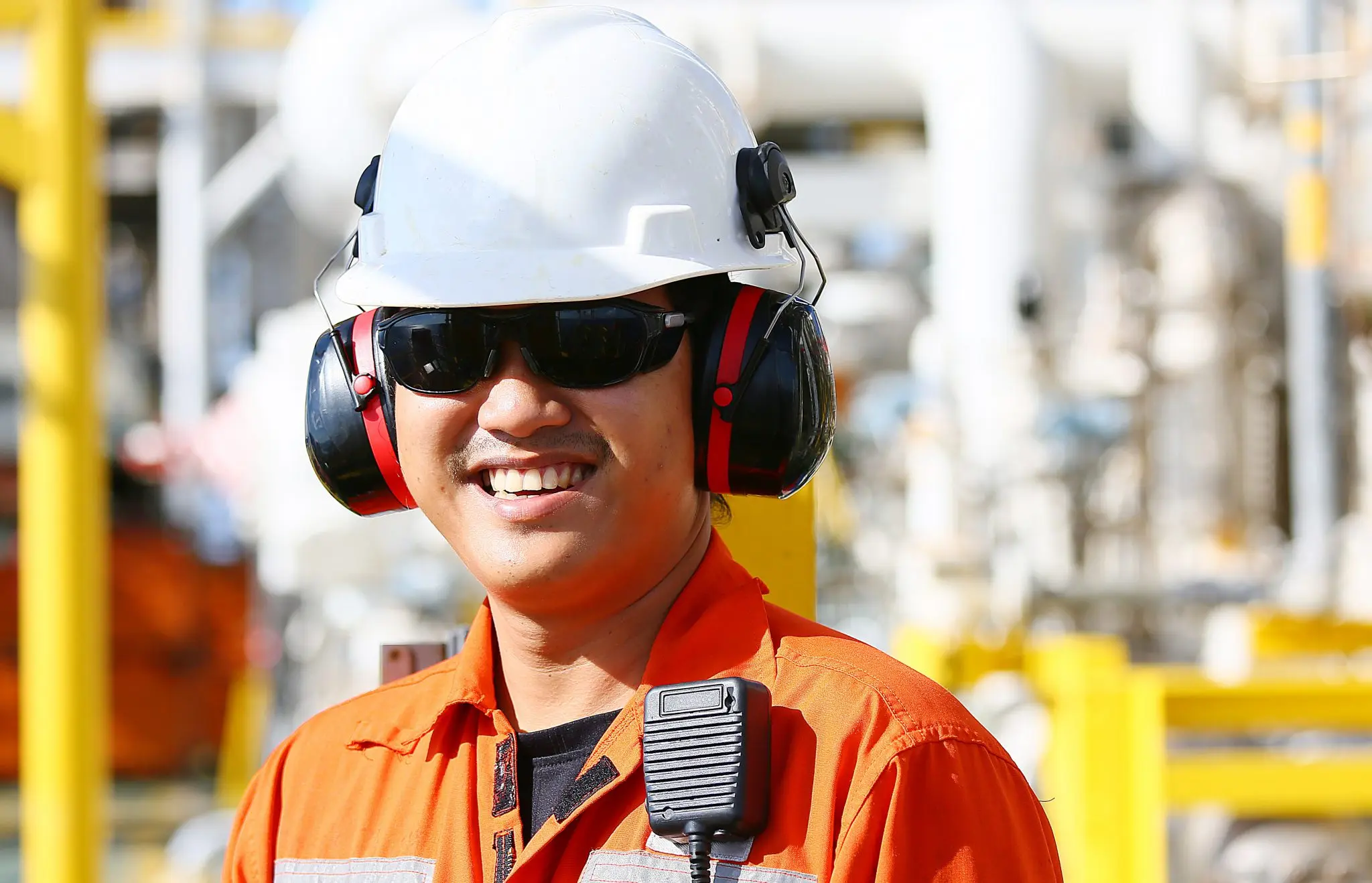 Safety officer wearing white hard hat, red and black ear protection, dark sunglasses, and orange safety vest with radio microphone on industrial work site