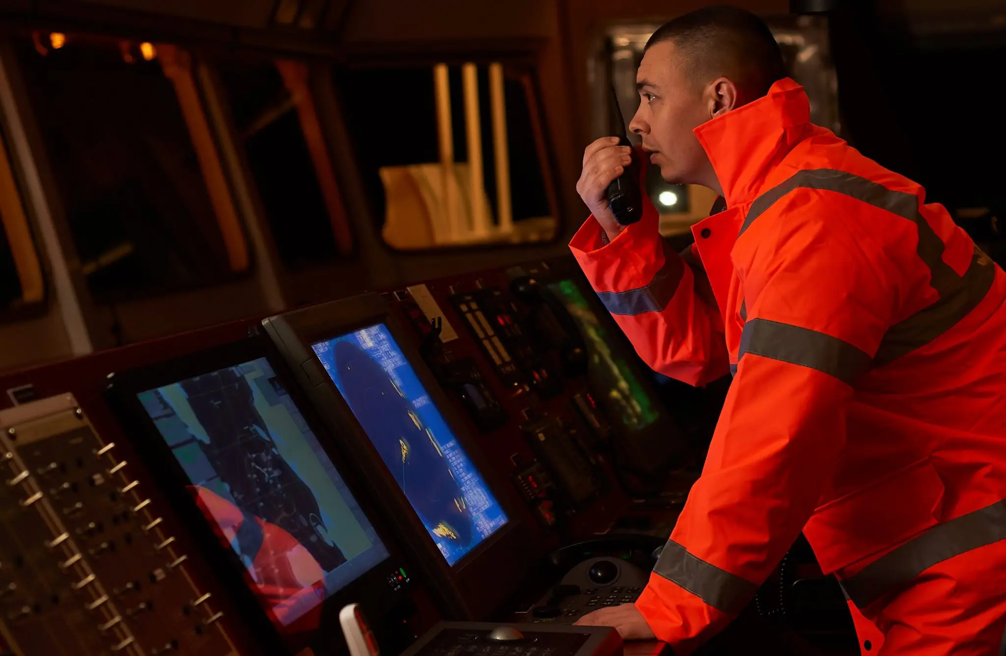 Maritime officer in red safety gear monitoring multiple screens displaying cyber security data on vessel bridge, demonstrating STCW cyber awareness training.