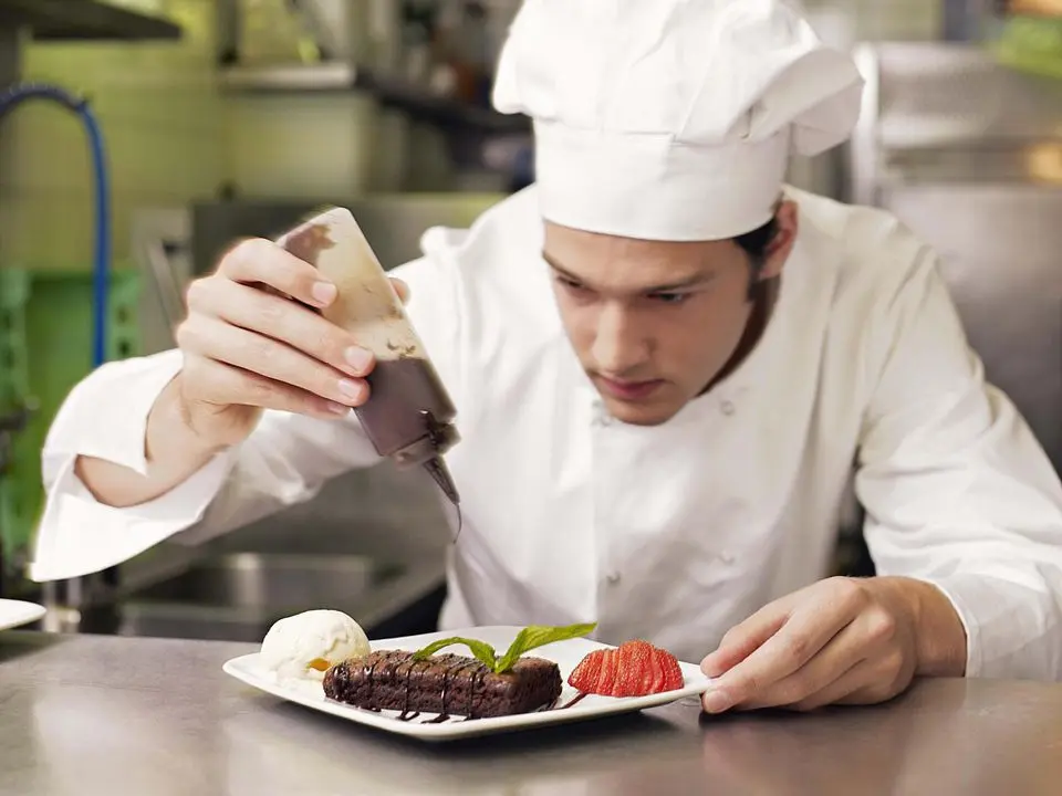 Professional chef in white uniform and hat carefully plating a gourmet dish with sauce, demonstrating food safety and presentation techniques for HACCP training