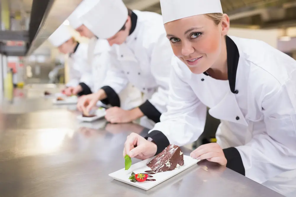Professional chefs in white uniforms and chef hats plating chocolate dessert with garnish in commercial kitchen, demonstrating food safety and hygiene practices for HACCP training