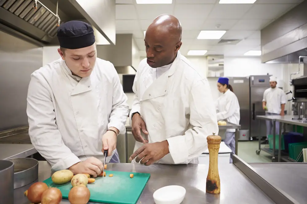 Two chefs in white coats demonstrating food safety and hygiene techniques by cutting vegetables in a professional kitchen during HACCP training.